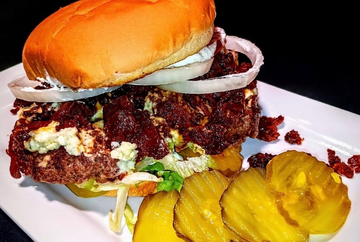 A burger topped with crumbled blue cheese and red onion on a white plate, with pickle slices alongside