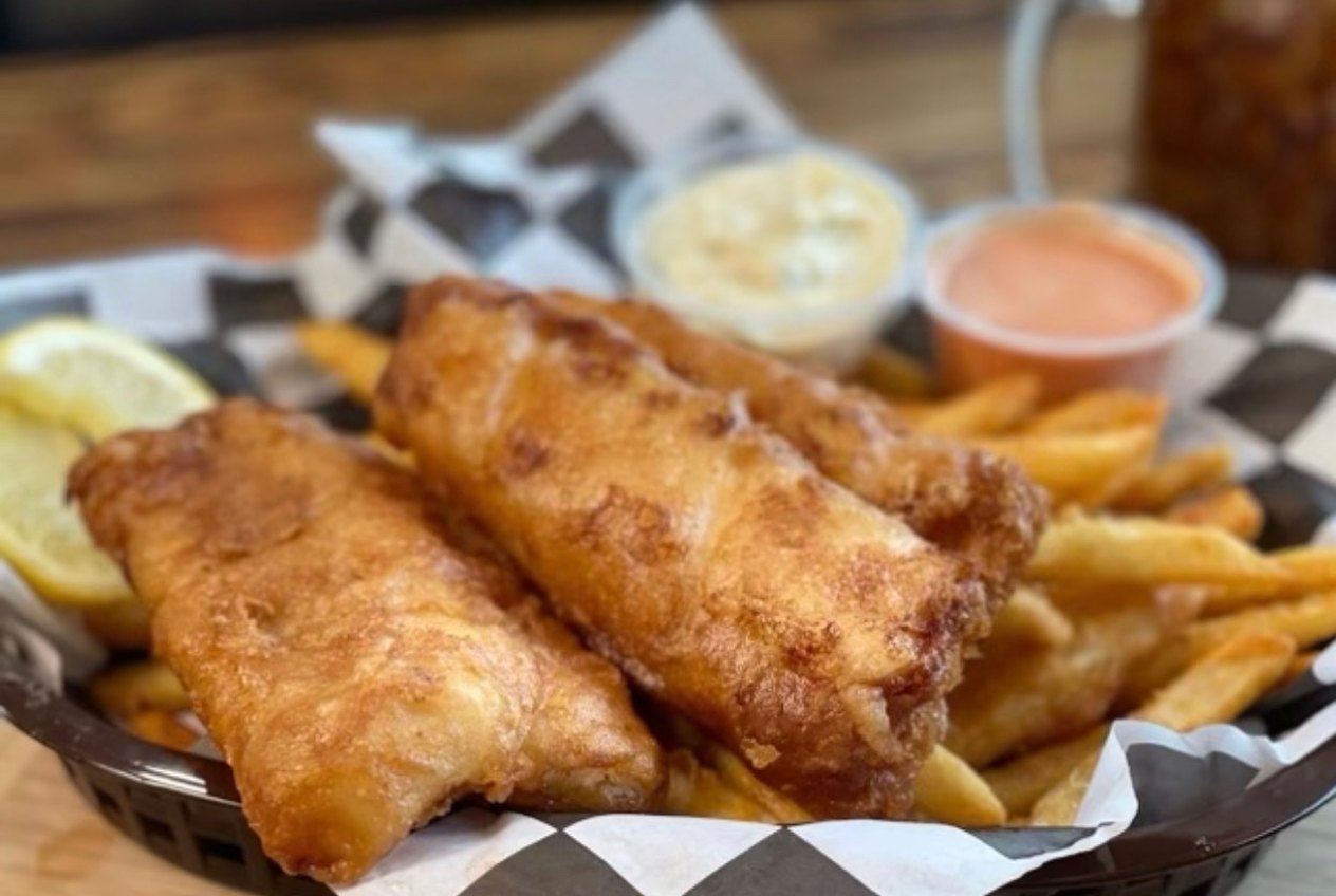 Two pieces of beer-battered cod in a paper-lined basket with fries, tartar sauce and a lemon wedge